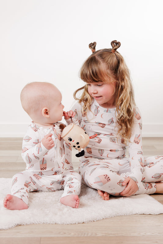 Two children in matching pajamas sitting on a white rug, holding a plush toy.