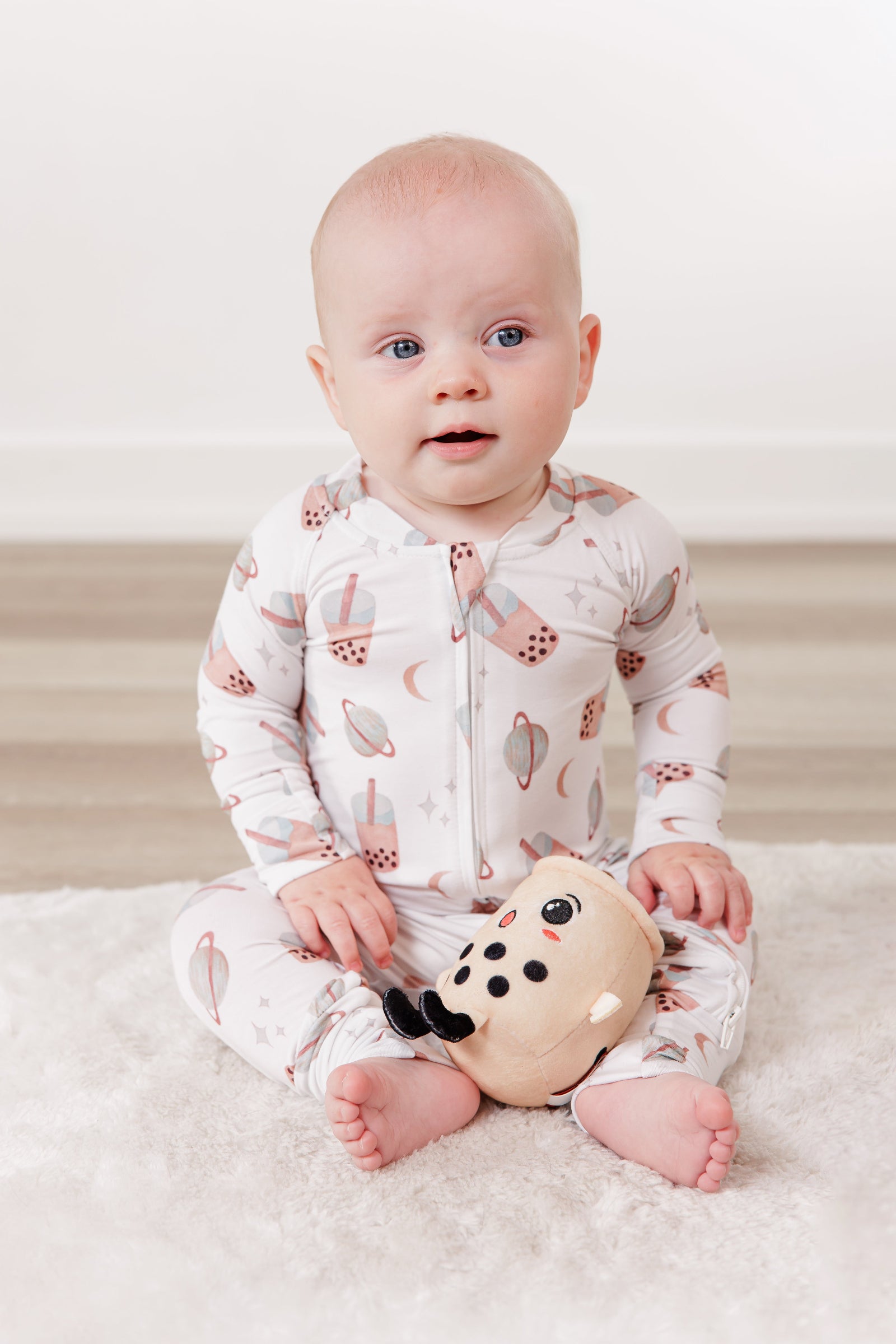 Baby in a patterned onesie holding a toy on a white blanket