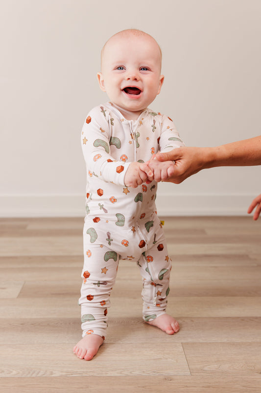 Baby in a gamer print onesie standing on a wooden floor with a person's hand holding their other hand.